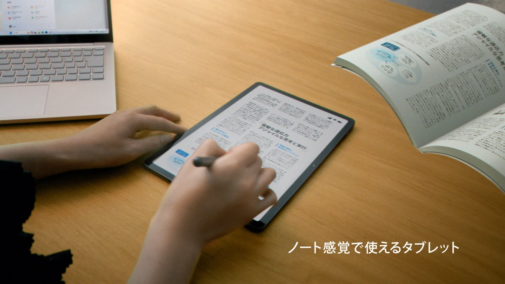 木製のテーブルの上にタブレット、本、ノートPCが置かれており、手でタブレットに書き込んでいる様子が写っています。タブレットには文章が表示されており、本も開かれています。