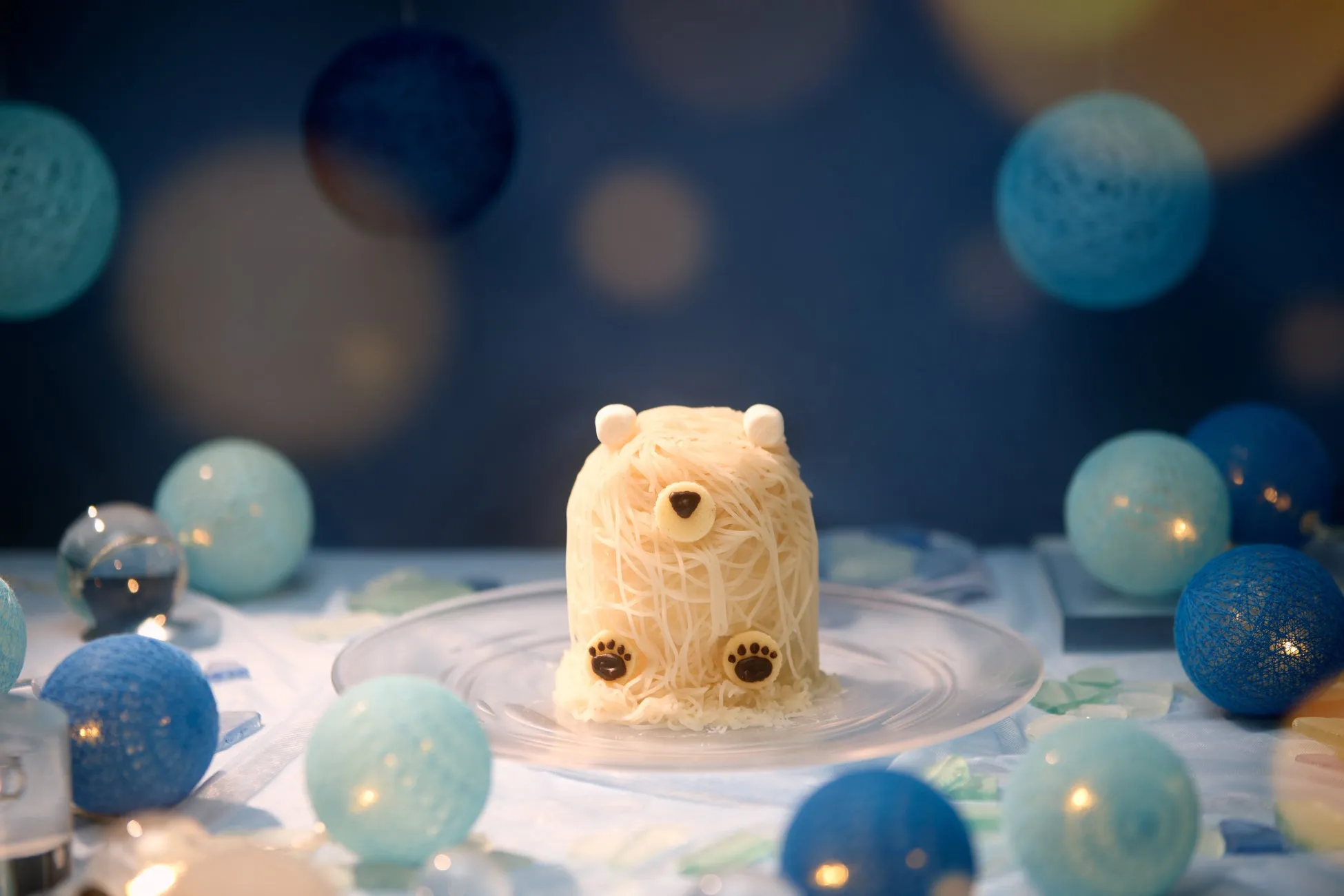 クマの形をしたデザートが皿に載せられ、周囲には青色の装飾品が配置されている写真です。背景は青色で、光が当たっています。