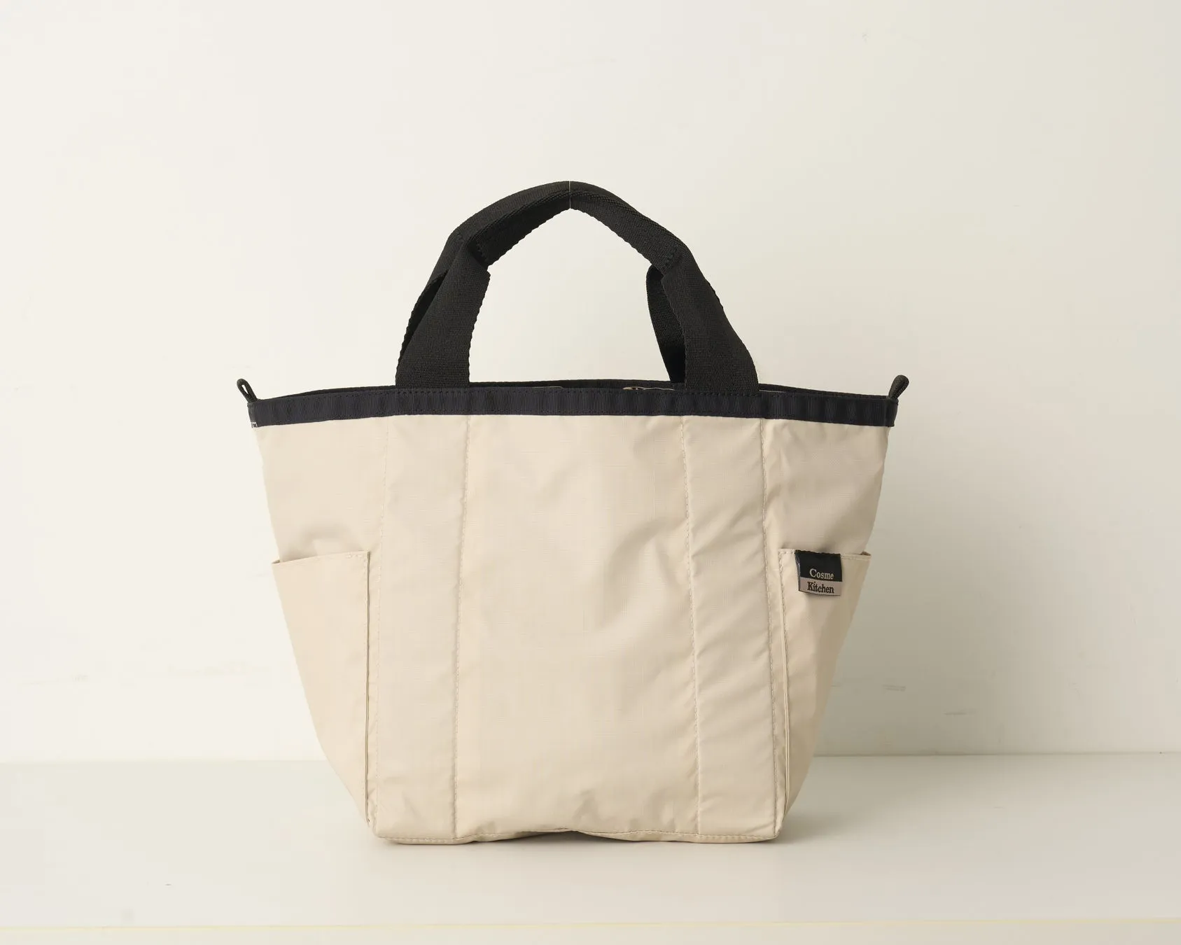ベージュ色のトートバッグの写真です。黒い持ち手と黒い縁取りが特徴的で、前面にはCosme Kitchenのロゴが入っています。