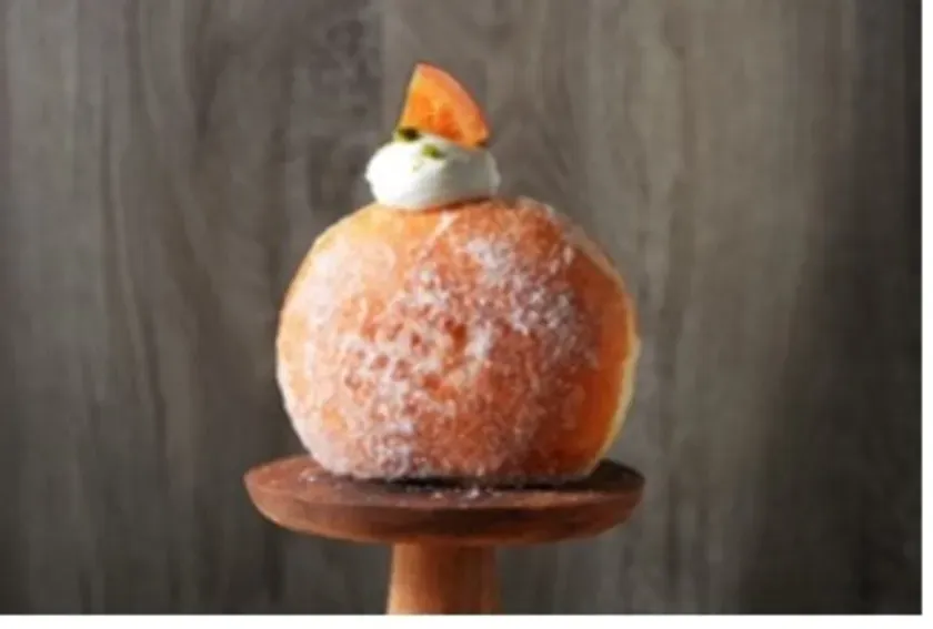 丸いドーナツが木製の台座に乗っている画像。ドーナツはオレンジ色で、粉砂糖がまぶされており、上に白いクリームとオレンジ色の飾りがある。