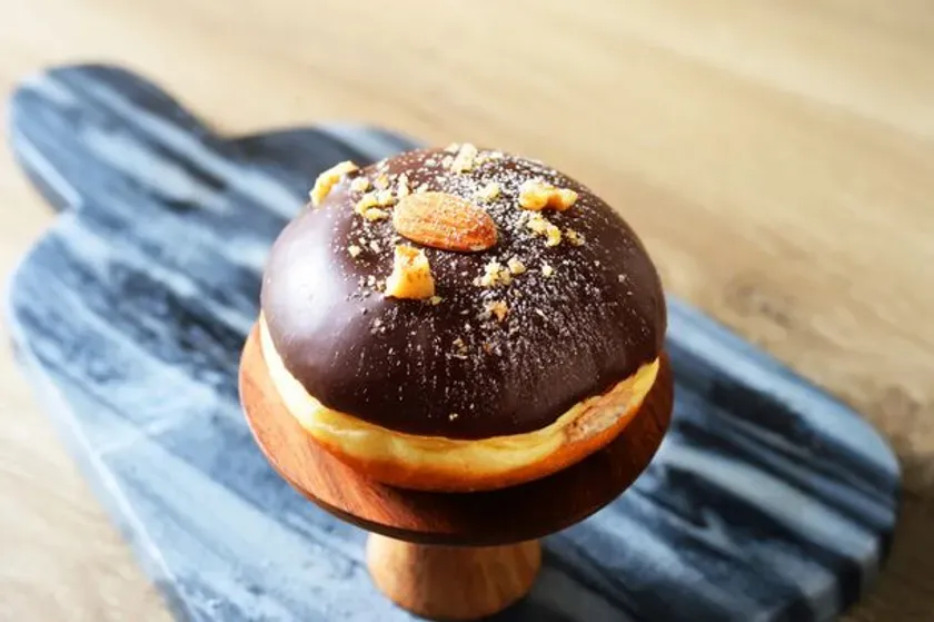 チョコレートでコーティングされ、アーモンドがトッピングされたドーナツが、木製の台座の上に置かれています。背景には木製のテーブルと大理石のボードがあります。