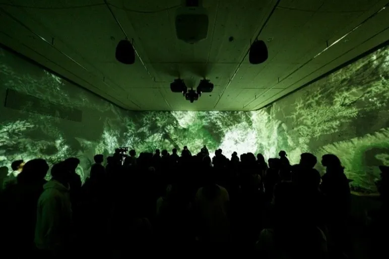 観客は、壁に投影された緑色の画像を見るために、暗い部屋に集まっている。天井にはプロジェクターとスピーカーが設置されている。