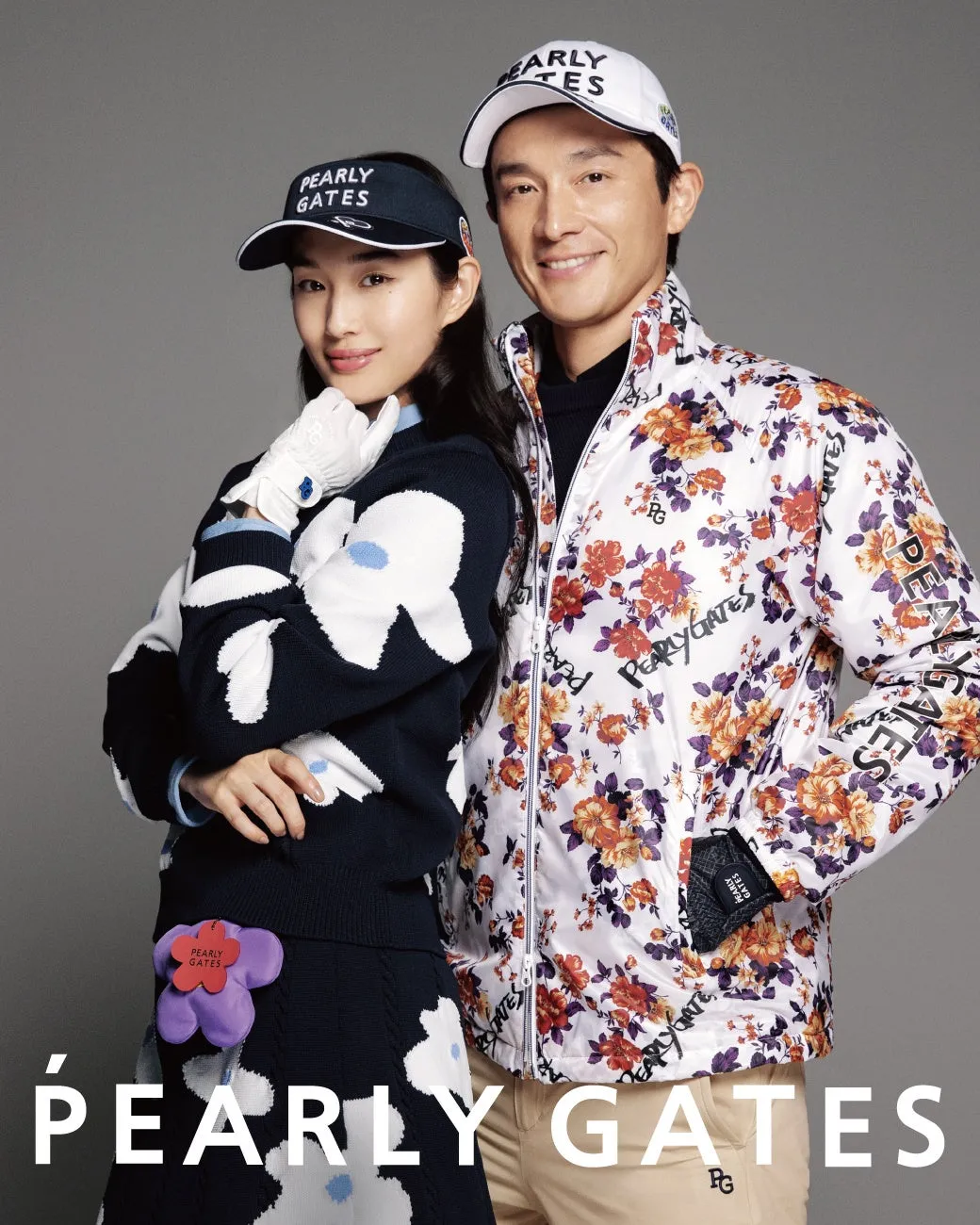 2人の人物が写っている写真。女性はネイビーのサンバイザーとセーターを着ており、男性は花の模様が入ったジャケットを着ている。背景はグレー。