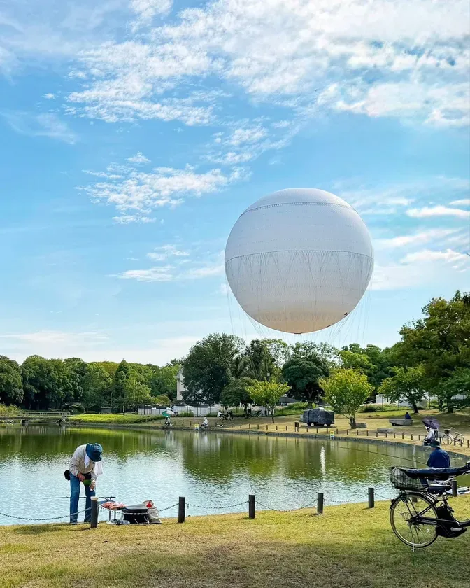 公園の池の上を巨大な白い風船が浮かんでいる様子を捉えた写真。池のそばでは、釣り人や自転車に乗った人が見られる。青空には雲が広がり、穏やかな晴天の下での風景である。