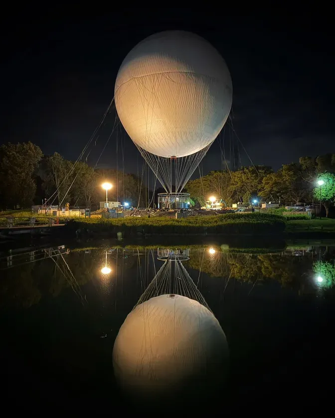 夜の湖に映る巨大な気球の様子を描いた写真。気球は水面に反射し、周囲には明かりが灯っている。