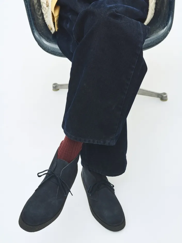 椅子に座っている人の足元の写真。濃い色のパンツ、赤色の靴下、スエードの靴が写っています。