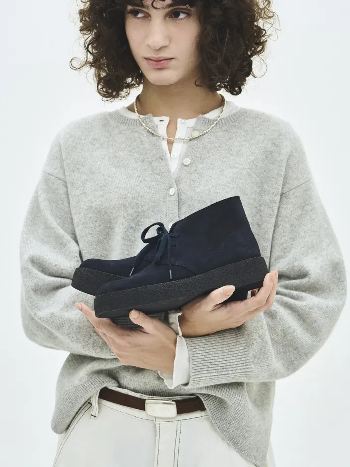 女性がネイビーのブーツを手に持っている様子を描いた写真。女性はグレーのカーディガン、白いシャツ、白いズボンを着用しており、茶色のベルトをしています。背景は白です。