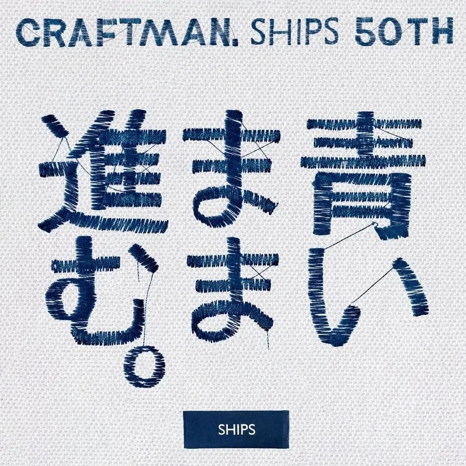藍色の刺繍で表現された日本語の文字と英字のロゴが特徴的な画像。背景は白い布地で、文字は「進む、お、ま、い」と読める。上部には「CRAFTMAN. SHIPS 50TH」と書かれており、下部には「SHIPS」のロゴが入っている。