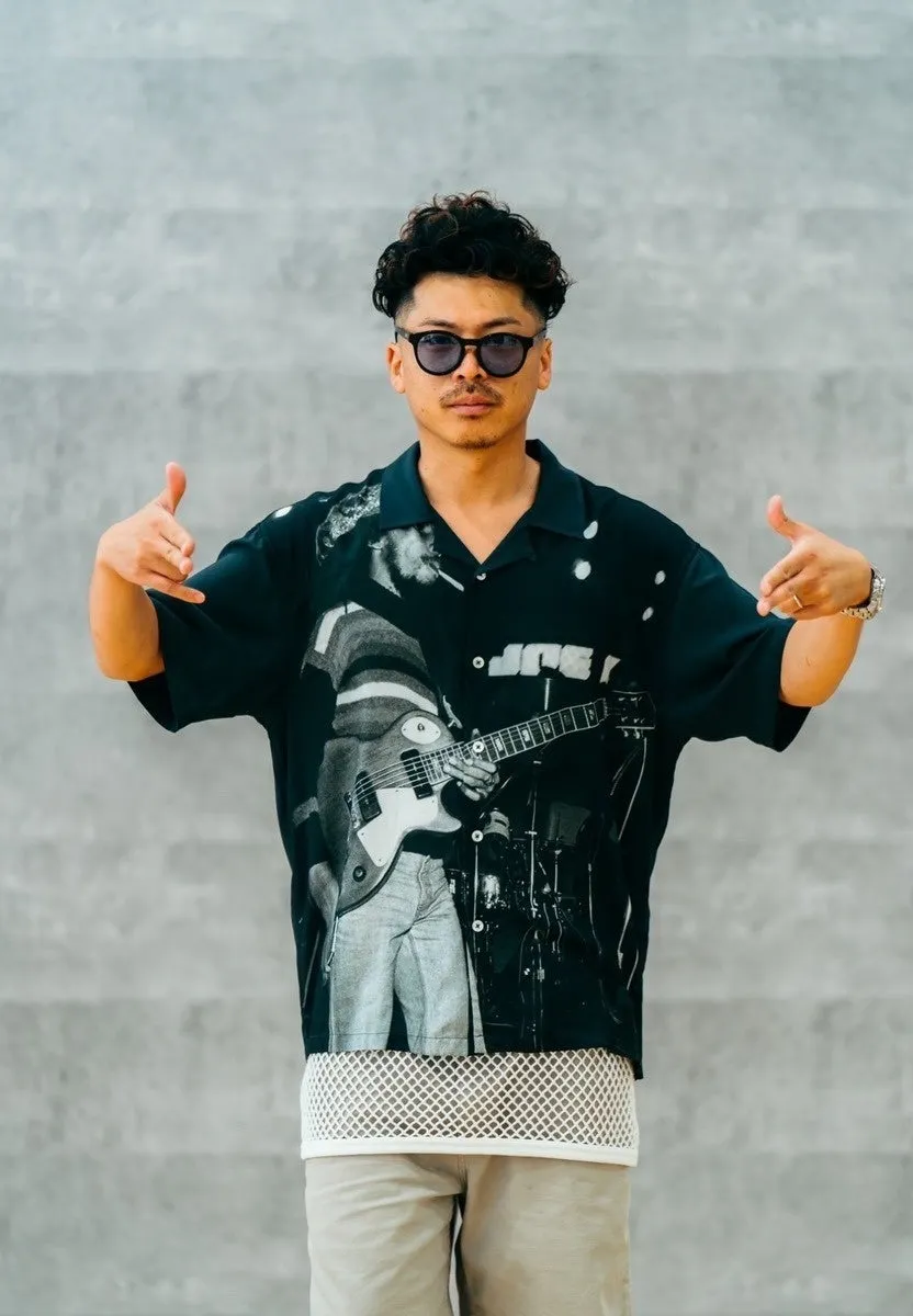 サングラスをかけた男性が、ギターが描かれた黒いシャツを着てポーズをとっている写真です。