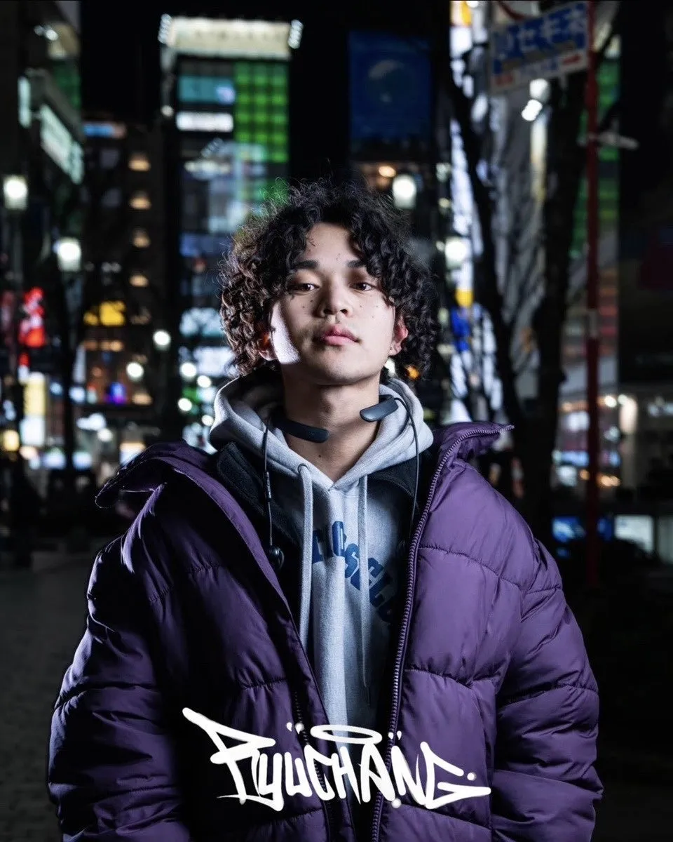 夜の街を背景に、紫色のダウンジャケットを着た男性が正面を見つめている写真です。彼の顔には光が当たり、前髪はカールしています。彼の首にはヘッドホンがあり、胸元にはロゴが描かれています。