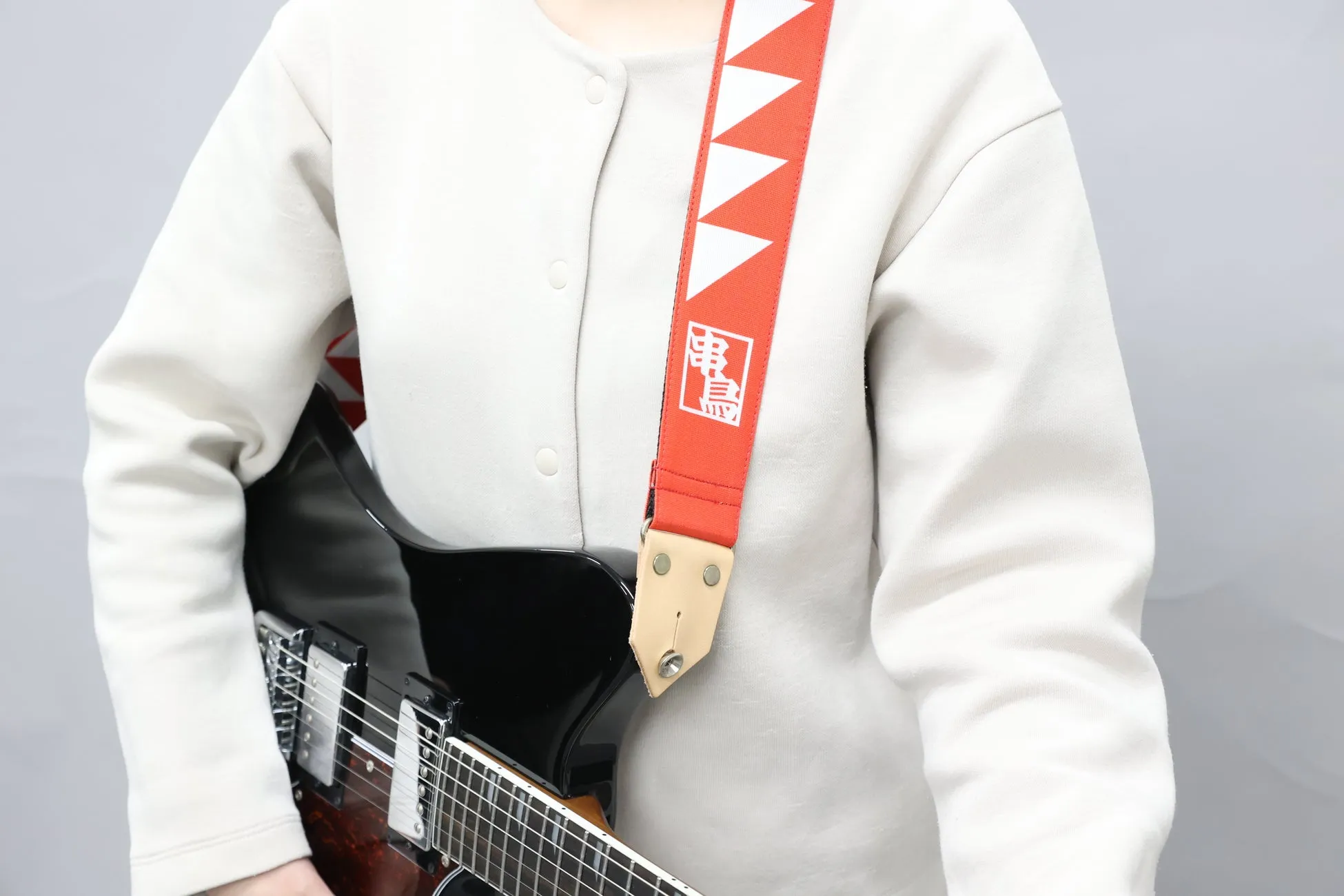 ギターを弾いている人の写真。赤いギターのストラップには、白い三角形と漢字が描かれています。白い服を着ています。