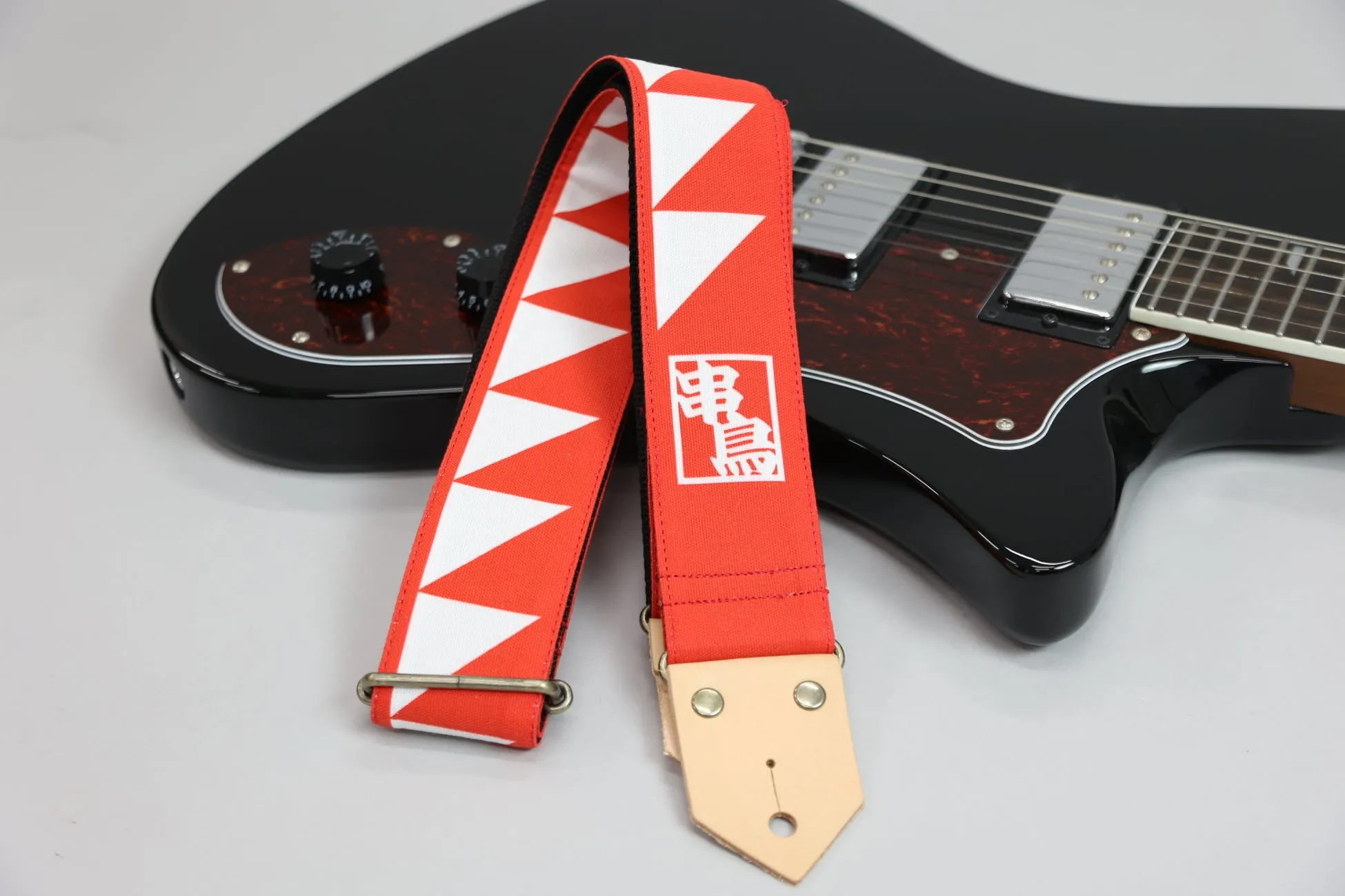 黒いエレキギターと、赤い三角模様のギター ストラップの写真です。ストラップには漢字の「電鳥」の文字が白抜きで入っています。