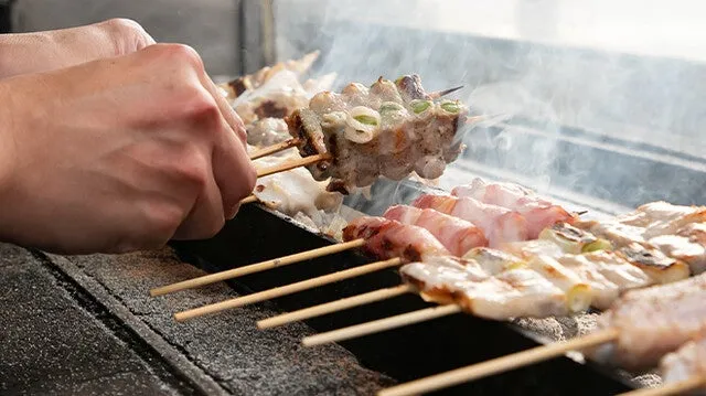 焼き鳥を焼いている様子を捉えた写真です。美味しそうな焼き鳥が炭火の上で焼かれており、手で串を持ちながら調理している様子が写っています。煙が立ち上り、食欲をそそる光景です。