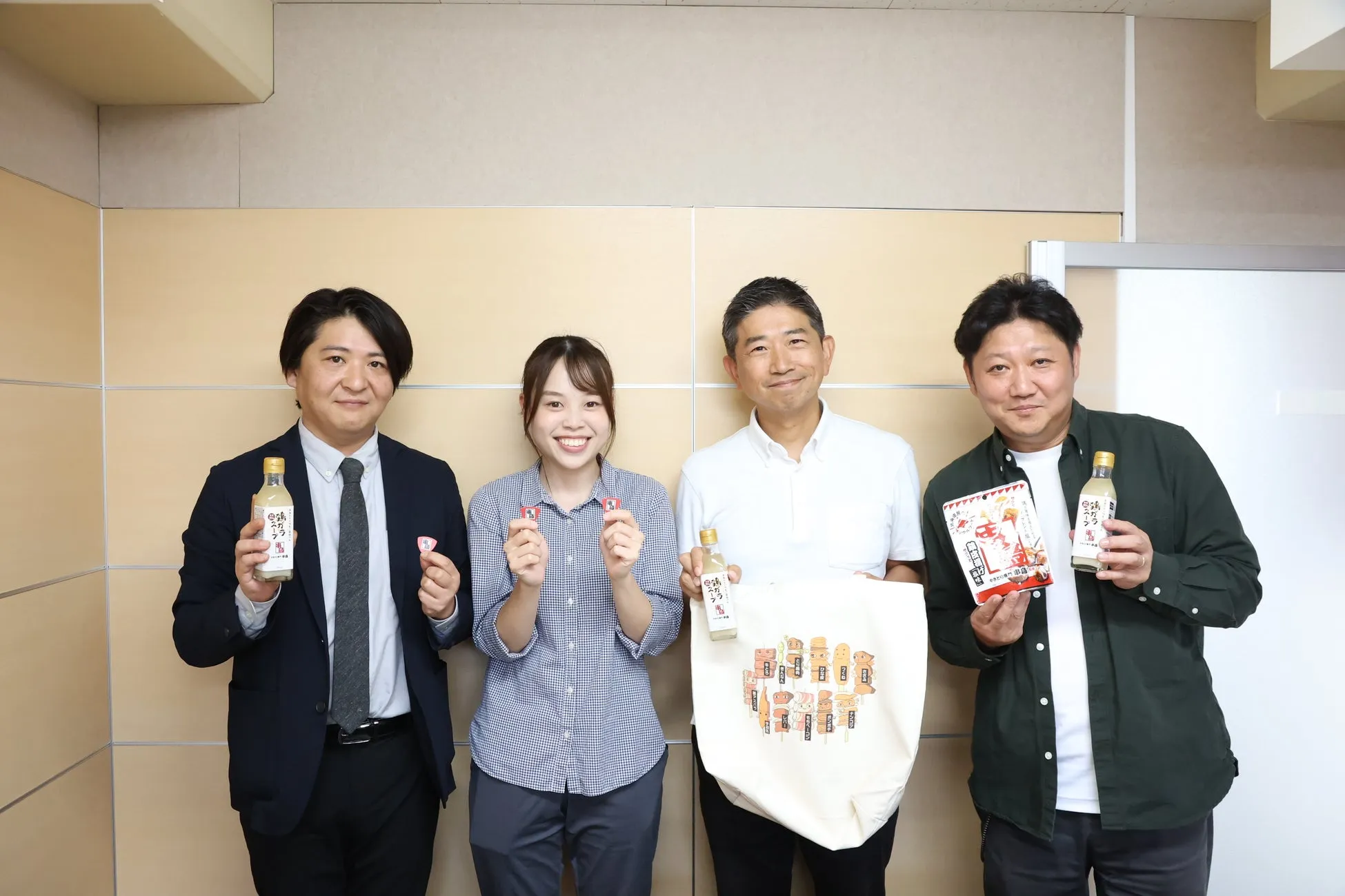 4人の人々が、製品を手に持って立っている写真。左の男性はボトルを、女性は小さなカードを2枚、真ん中の男性はトートバッグとボトルを、右の男性は箱とボトルを持っています。彼らは笑顔でカメラを見ています。