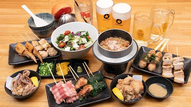 テーブルの上に、様々な料理が並べられています。焼き鳥、サラダ、ご飯、揚げ物、ビールなどがあり、居酒屋の食事風景を捉えた写真です。