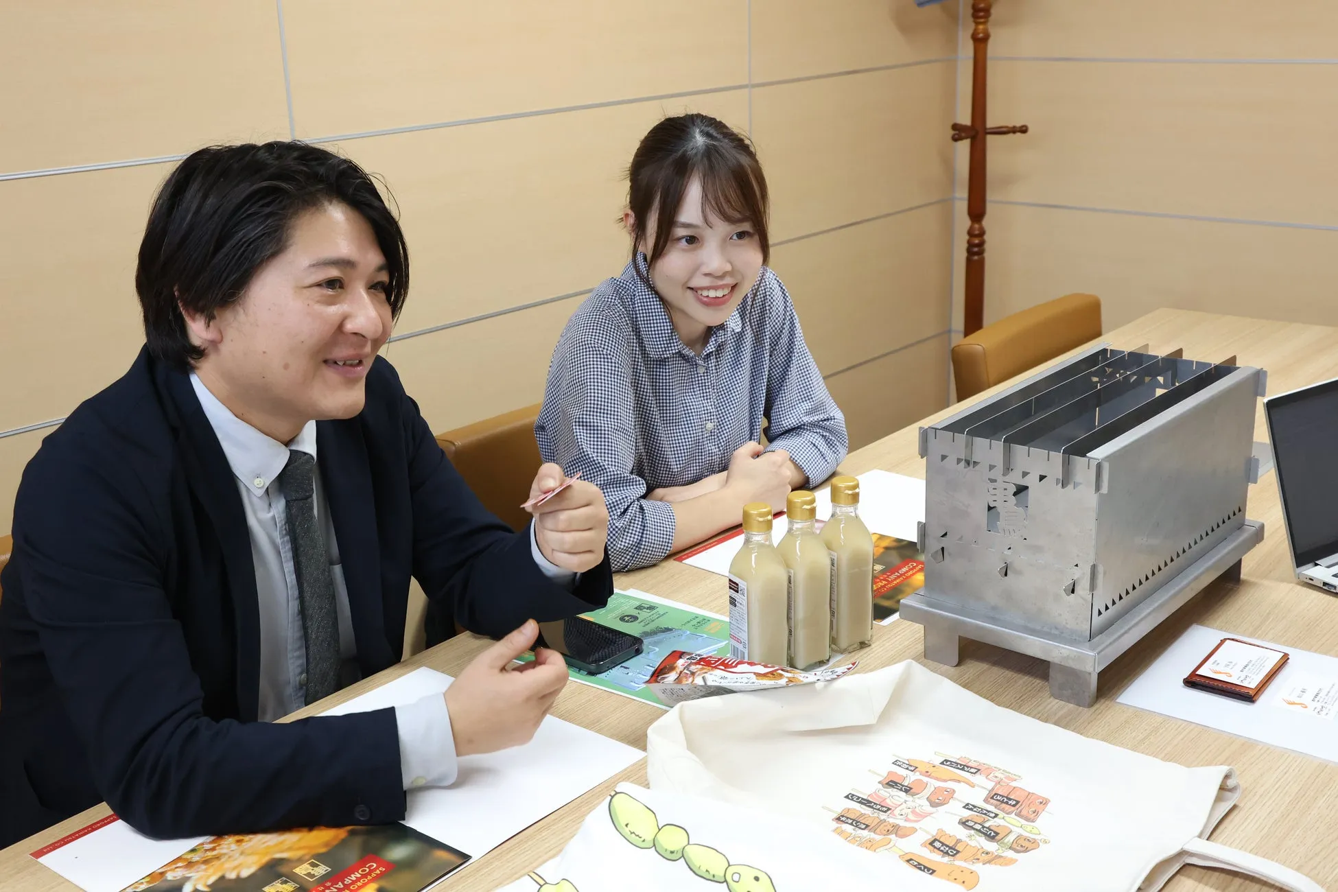 会議室で、男女2人が笑顔で話している様子。テーブルには、焼き鳥用のコンロ、飲み物、トートバッグなどが置かれている。