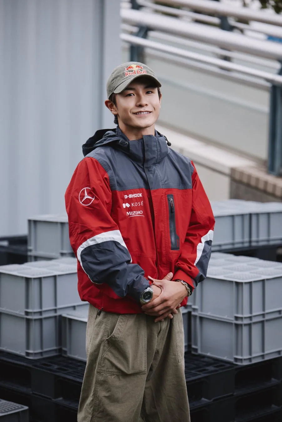 赤いジャケットを着て、Red Bullの帽子をかぶった男性が、笑顔で立っている写真です。背景には、積み重ねられたグレーの容器があります。