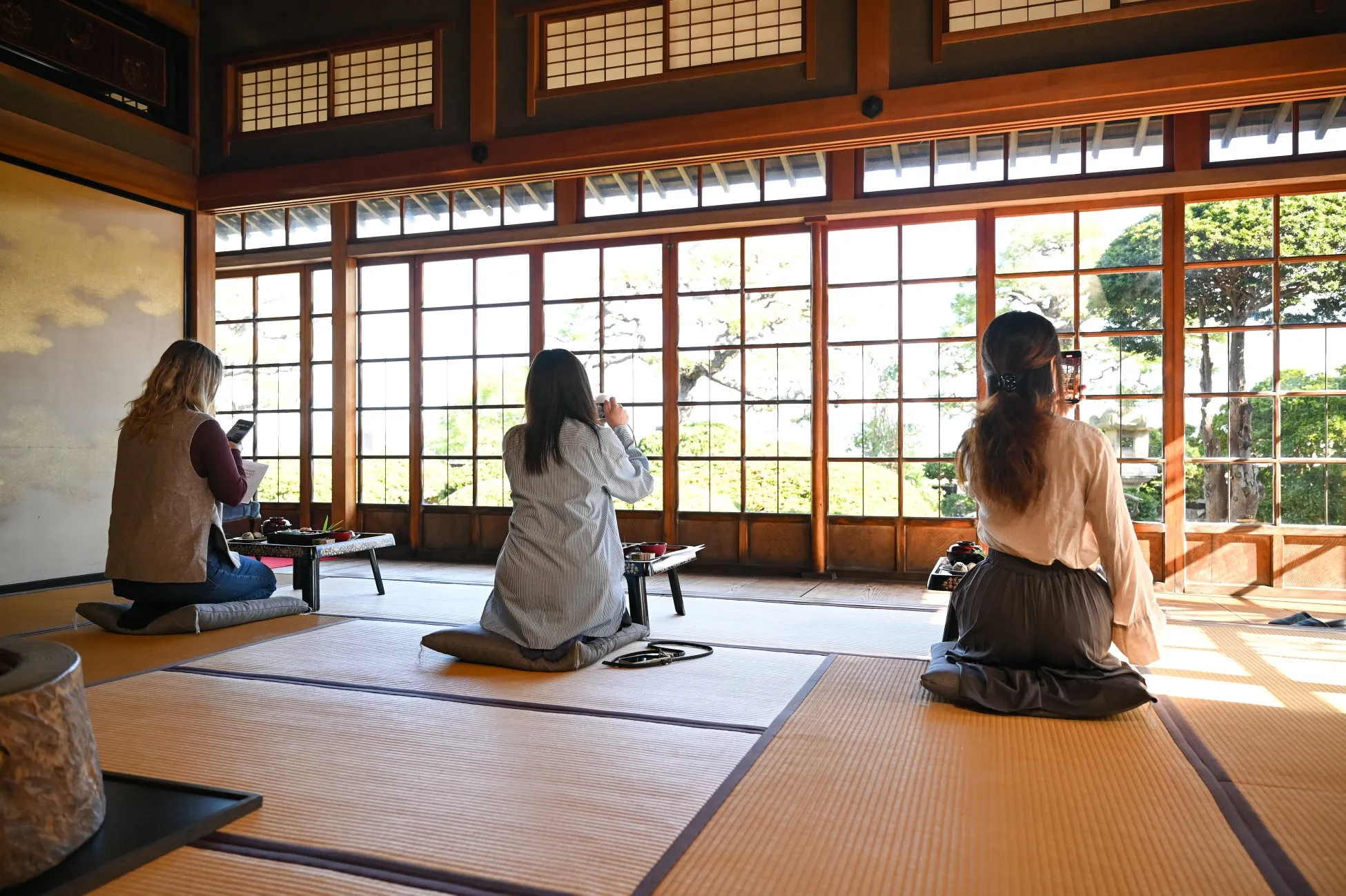 日本家屋と思われる部屋で、3人の女性がそれぞれ座布団に座り、庭を眺めたり、スマートフォンで写真を撮ったりしている様子