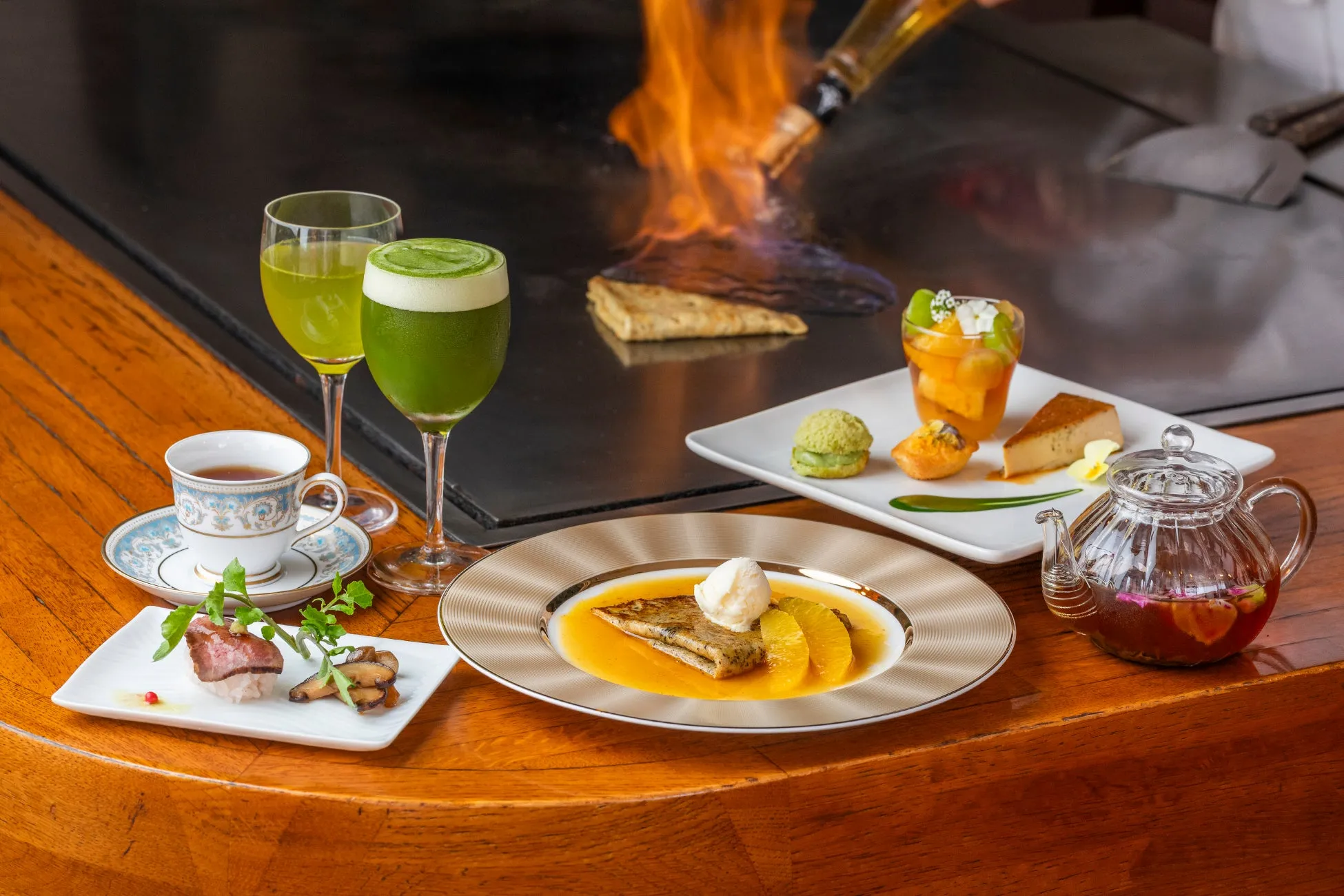 鉄板焼きレストランで撮影された、コース料理の写真です。焼かれた料理、デザート、飲み物、お茶のセットがテーブルに並んでいます。シェフが料理に火をつけている様子も写っています。