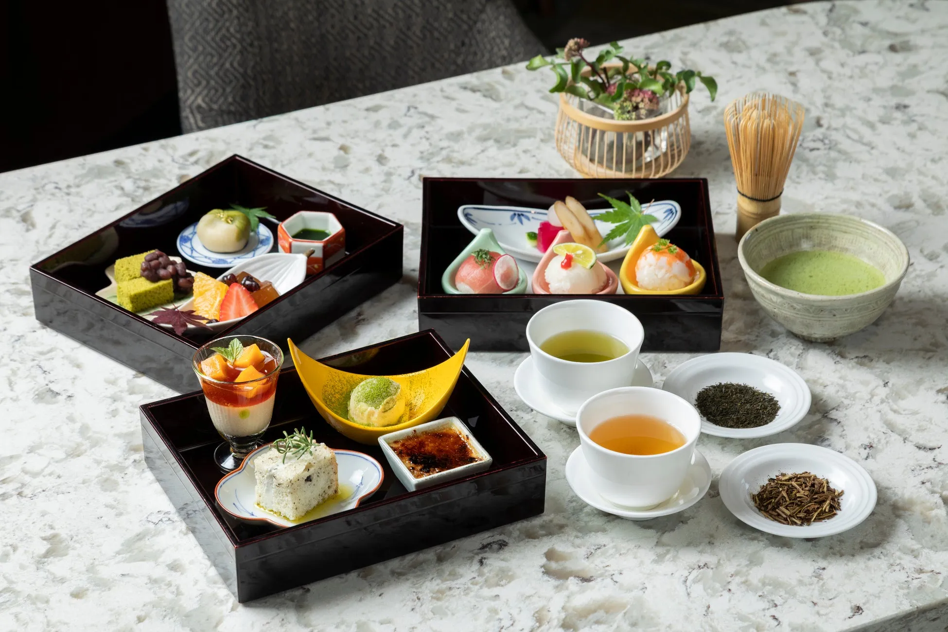 テーブルの上に置かれた、日本料理のセットのクローズアップ。セットには、様々な種類の料理を盛り付けた黒いトレイ、抹茶、お茶、茶葉が含まれています。