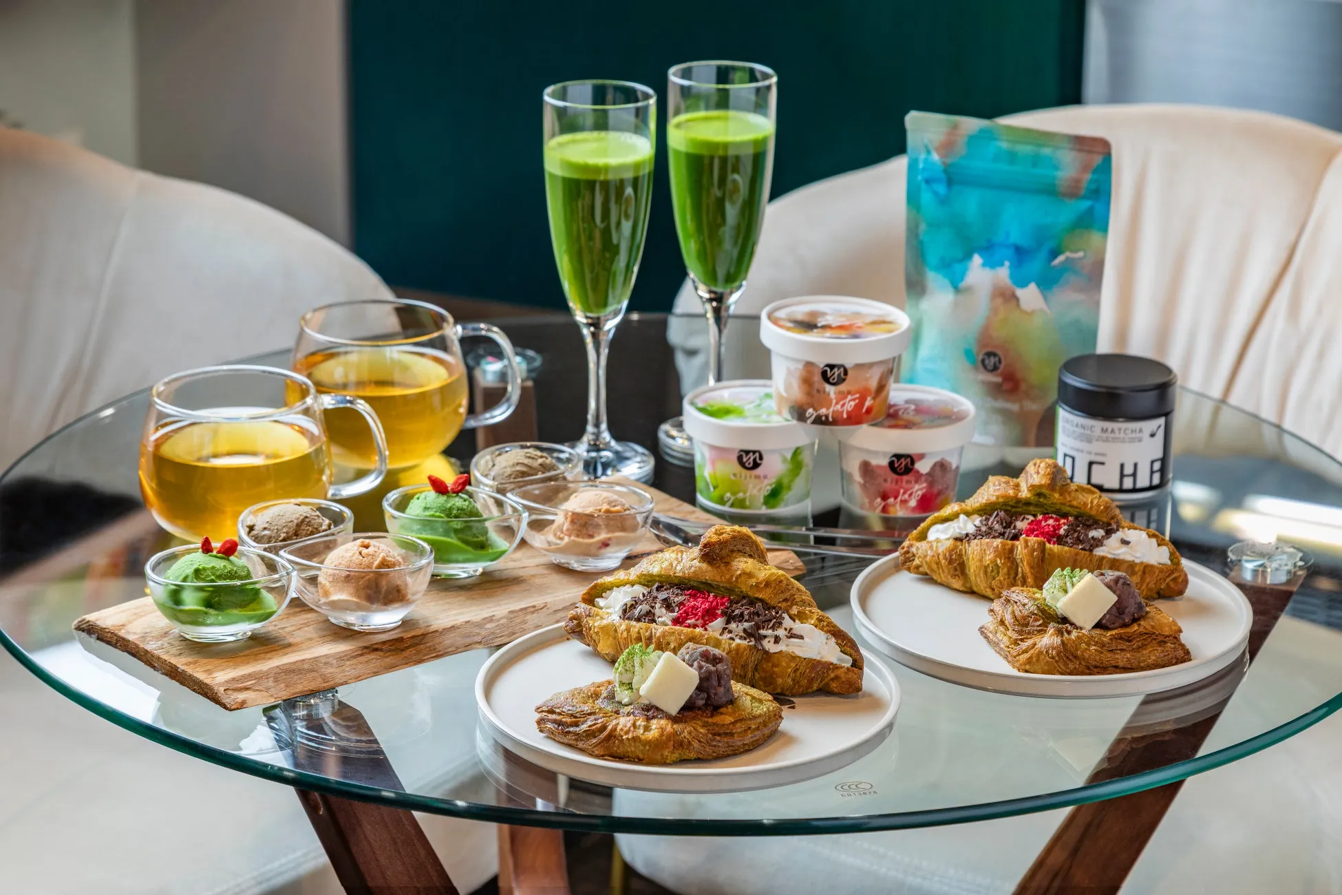 テーブルの上に、クロワッサン、アイスクリーム、抹茶、お茶、ゼリーなどの様々な食べ物と飲み物が並べられた写真です。アイスクリームは様々なフレーバーがあり、クロワッサンにはクリームやトッピングが添えられています。抹茶とゼリーはグラスに入れられ、お茶はティーカップに注がれています。すべてが洗練された雰囲気で配置されており、デザートや軽食のセットとして提供されているようです。