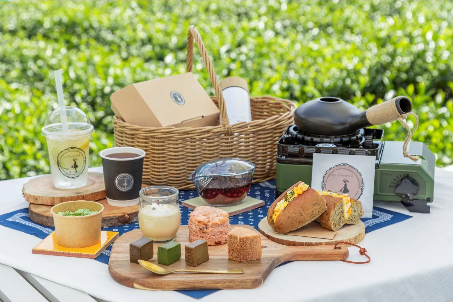 ピクニックのテーブルに、様々な食べ物と飲み物が並べられた写真。バスケット、飲み物、サンドイッチ、焼き菓子などがあり、屋外での食事シーンを演出している。