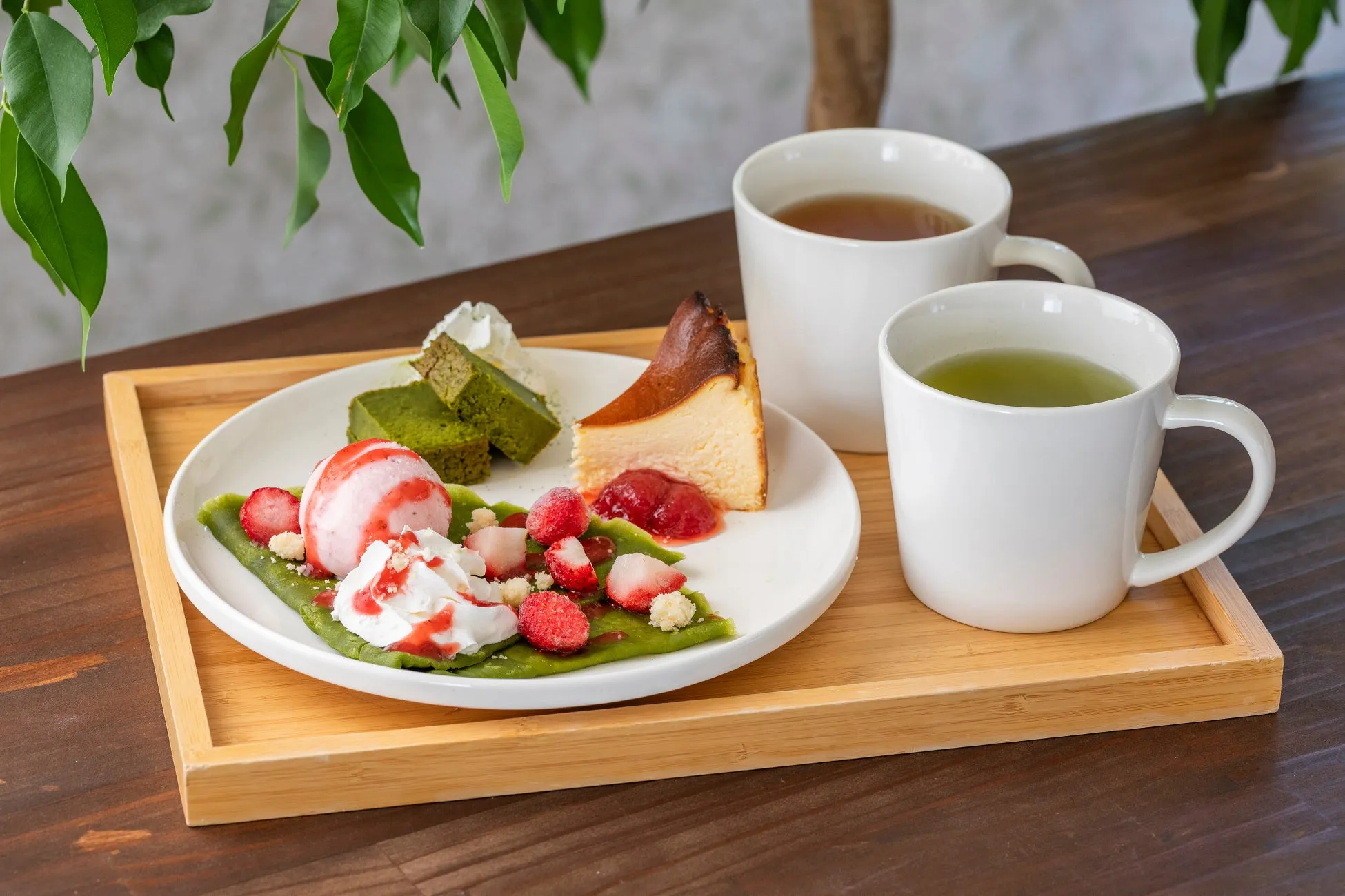 木製のトレイに、抹茶ケーキ、チーズケーキ、ストロベリーアイス、生クリーム、イチゴ、お茶が入った白いマグカップ2つが置かれている写真です。背景には緑の葉が見えます。