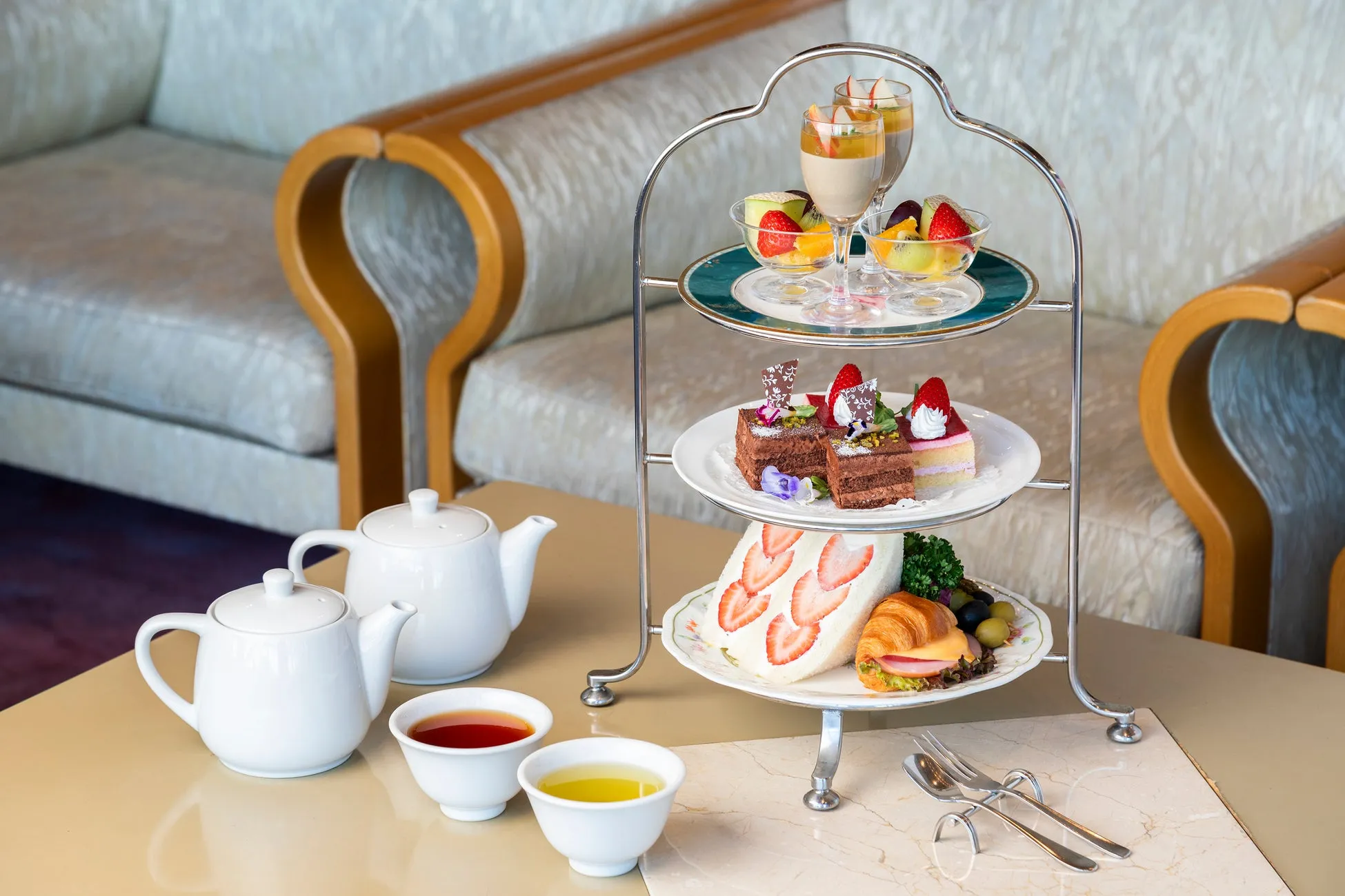 アフタヌーンティーのセットがテーブルに置かれている写真です。三段のスタンドには、ケーキ、サンドイッチ、フルーツ、ゼリーなどが並び、白いティーポットとカップには紅茶が注がれています。
