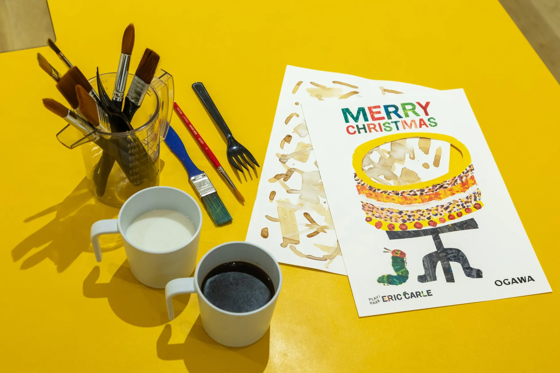 クリスマスをテーマにしたイラストと、絵筆、カップ、フォークなどの画材が配置された写真。