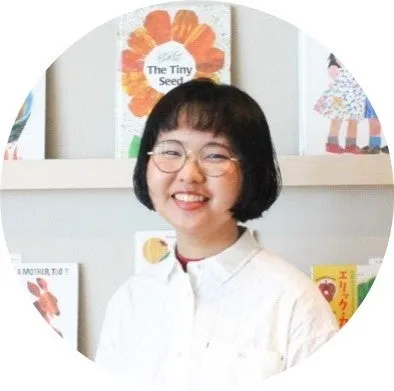 眼鏡をかけた女性が笑顔で本棚の前に立っている写真。