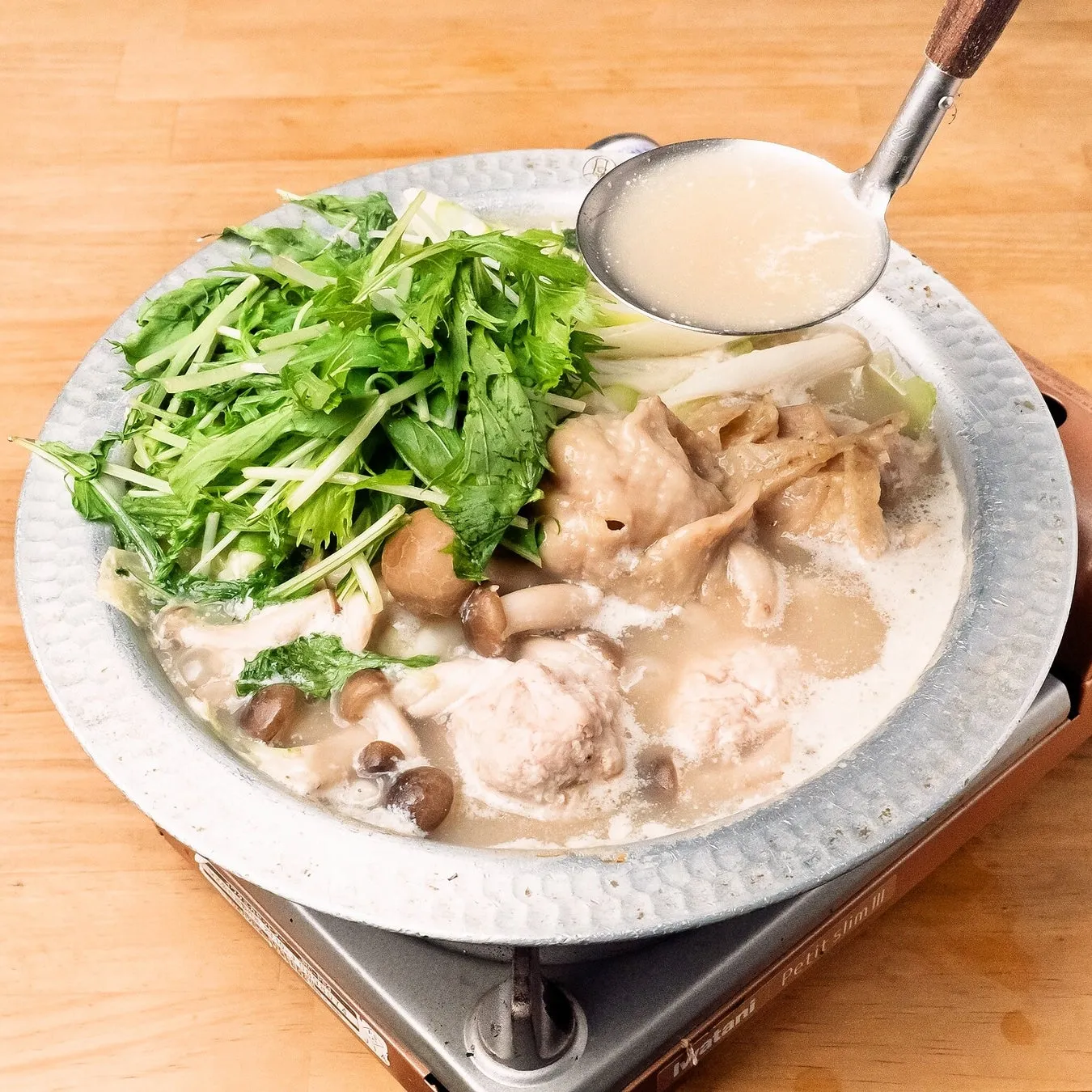 鍋料理の写真。具材は鶏肉、きのこ、野菜など。お玉でスープをすくっている様子。