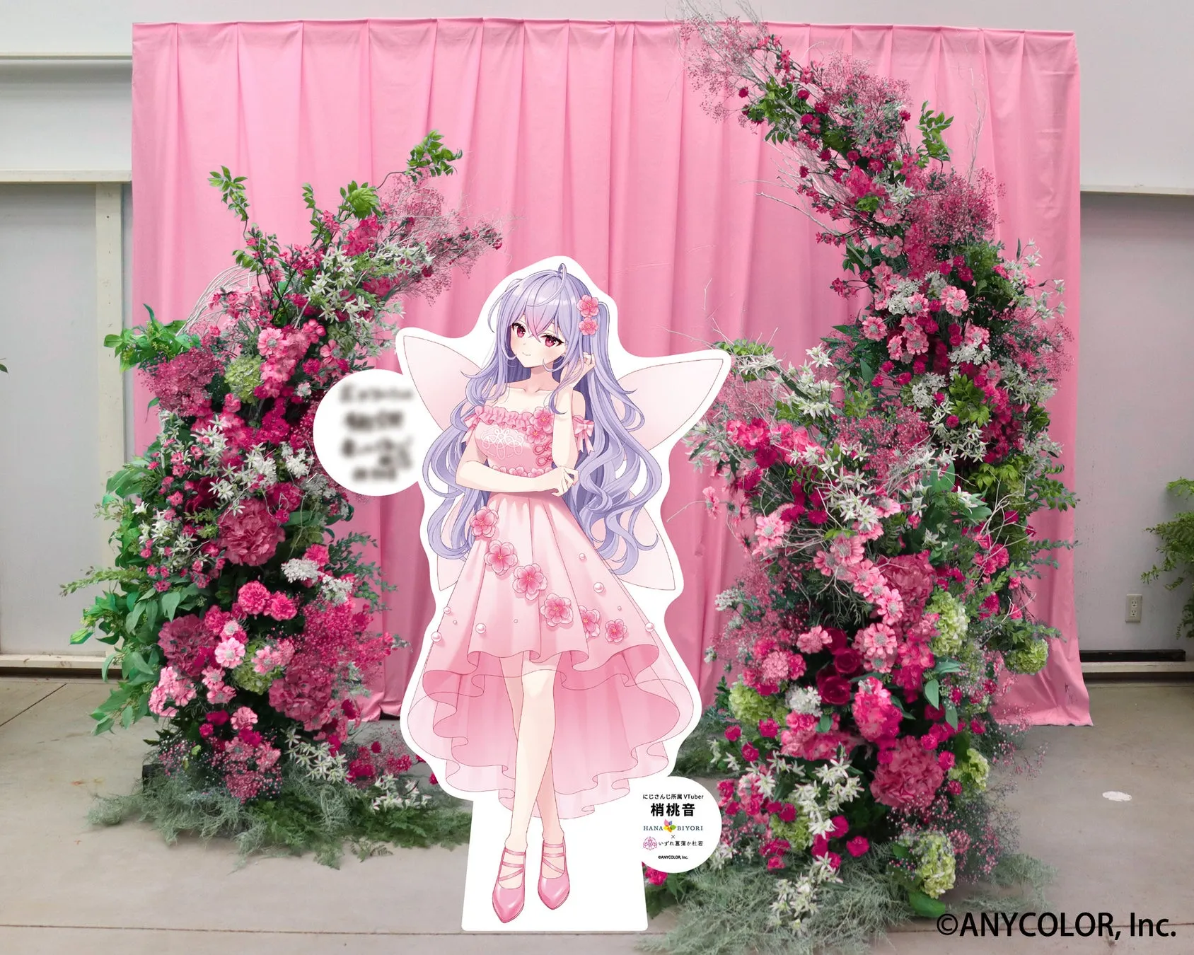 ピンク色の背景に、花で飾られたアーチとVtuberの梢桃音の等身大パネル。