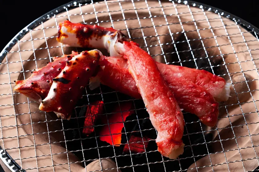 焼き網の上で焼かれているカニの足のクローズアップ写真。炭火の熱で赤く染まったカニの足が美味しそう。