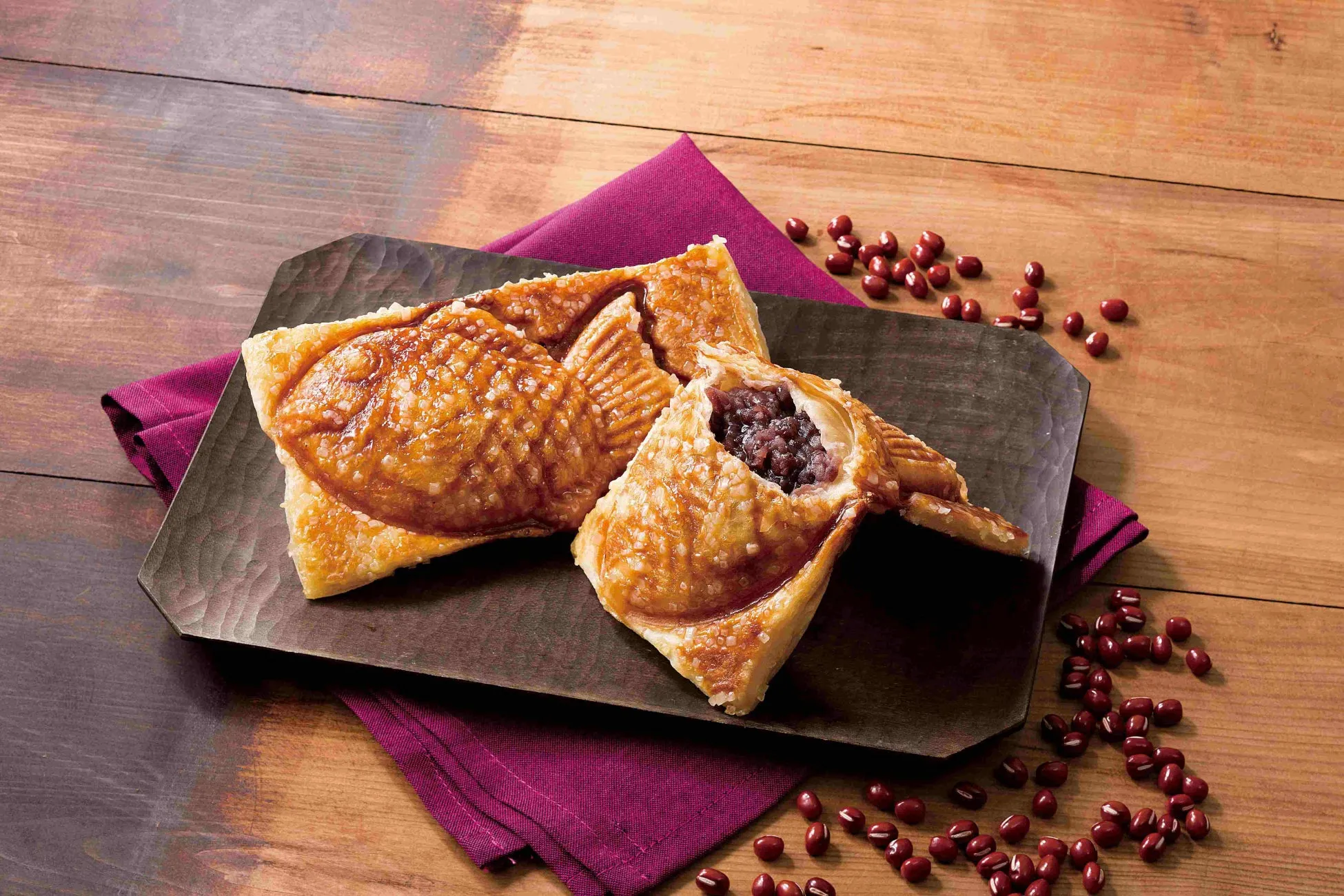 たい焼きが2つ、木製のプレートに乗せられており、そのうちの1つは餡が見えるように割れている。周りには小豆が散りばめられ、紫色の布が添えられている。