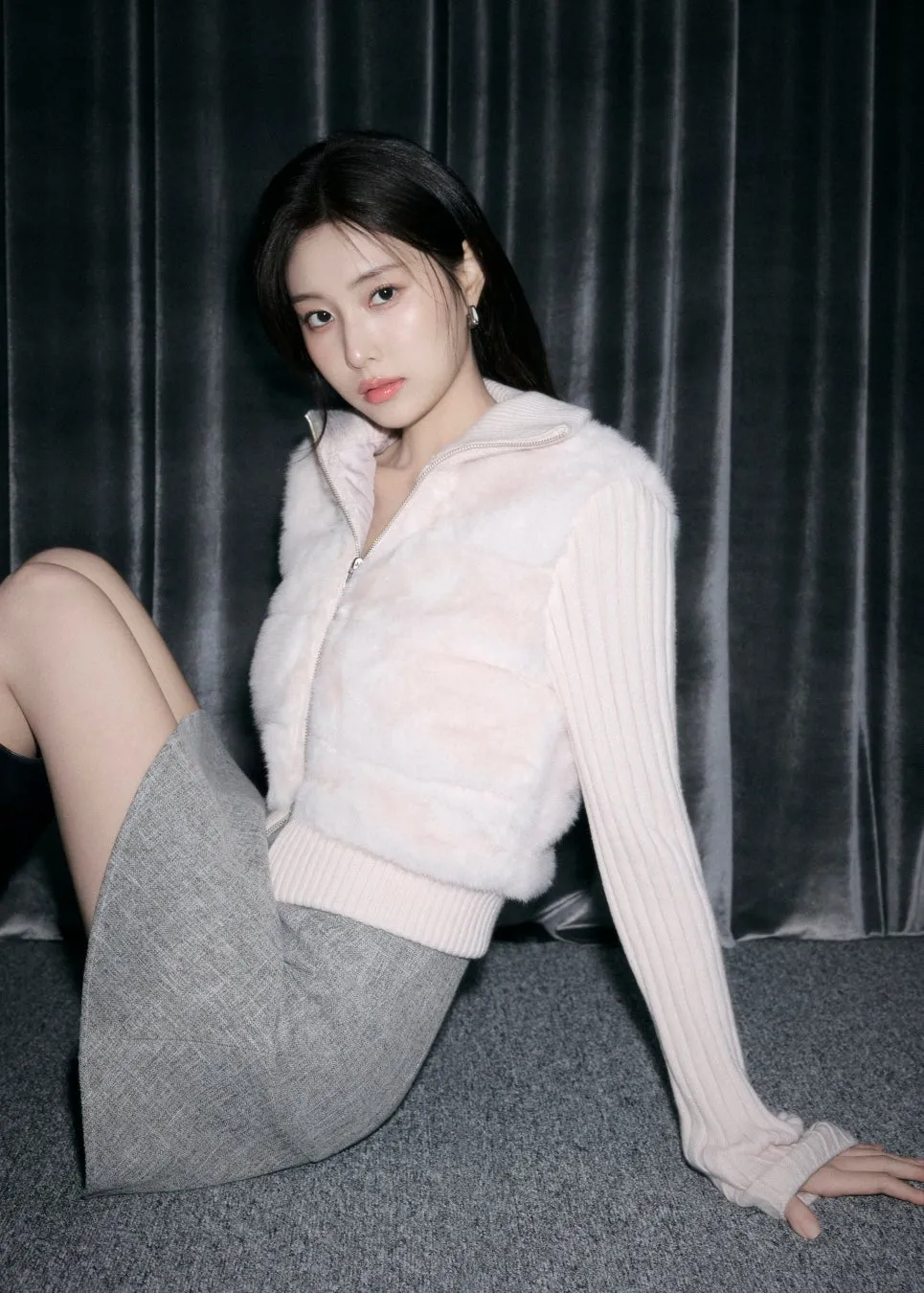 女性が座ってカメラを見つめている写真。彼女は白い毛皮のベストと灰色のスカートを着ており、背景は暗いカーテンです。