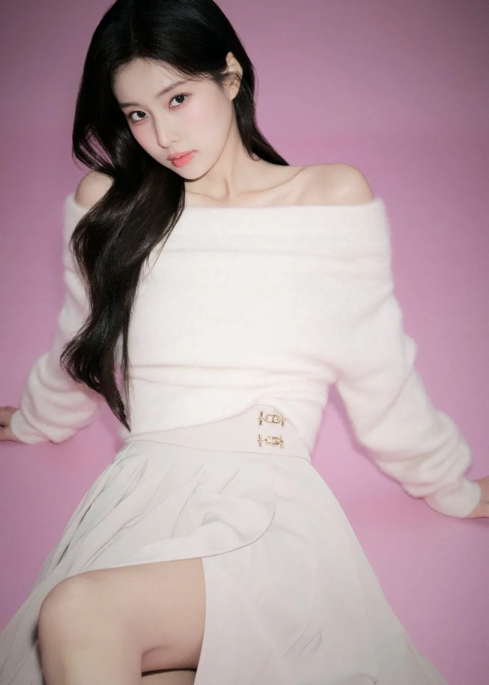 ピンクの背景にオフショルダーの白いセーターとスカートを着た女性が座っている写真です。