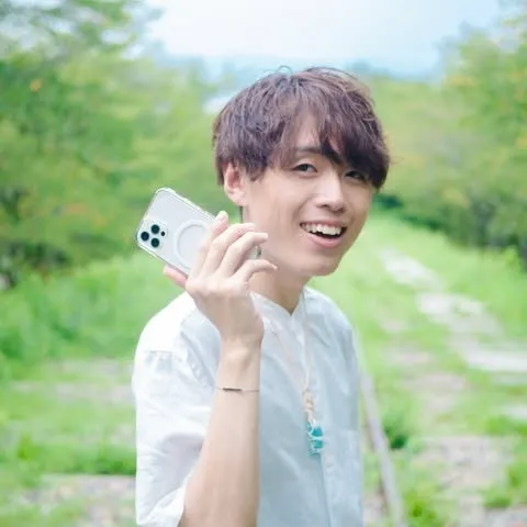 笑顔の男性がスマートフォンを持ち、屋外でポーズをとっている様子を捉えた写真です。背景には緑色の木々や草木があり、自然光が差し込んでいます。男性は白いシャツを着ており、カジュアルな印象を与えます。