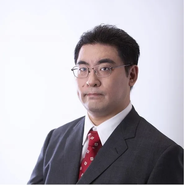 眼鏡をかけた男性のポートレート。彼はグレーのスーツと赤いネクタイを着て、白い背景の前に立っています。