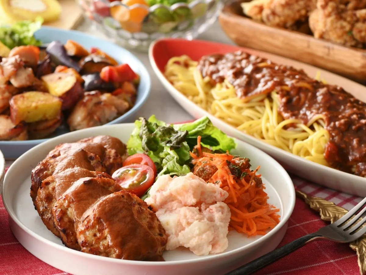 様々な料理がテーブルに並べられた写真。ハンバーグ、ミートソースパスタ、サラダ、揚げ物、野菜の煮物など、様々な料理が写っている。