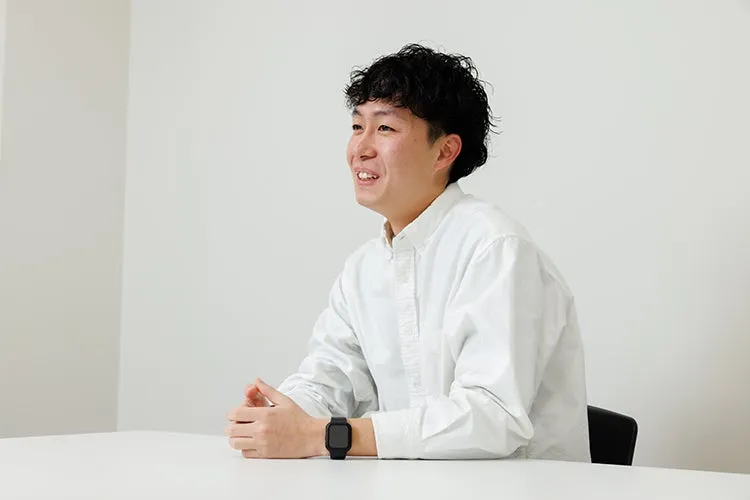 白いシャツを着た笑顔の男性がテーブルに座っている写真です。彼は黒いスマートウォッチを着用しています。