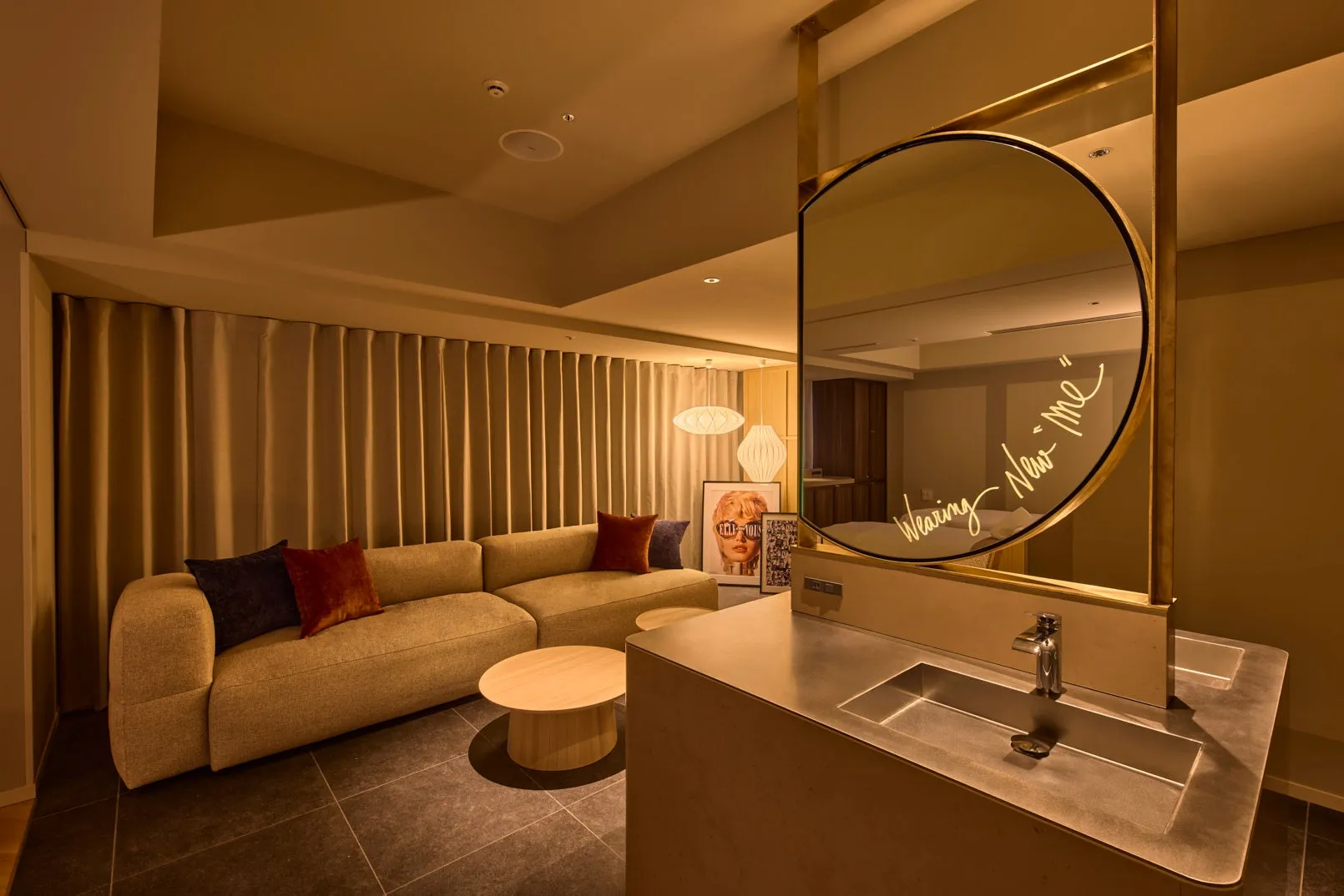 モダンな内装の部屋。丸い鏡があり、「Wearing New me」という文字が書かれている。ソファ、テーブル、照明、洗面台など、洗練されたデザインの家具が配置されている。