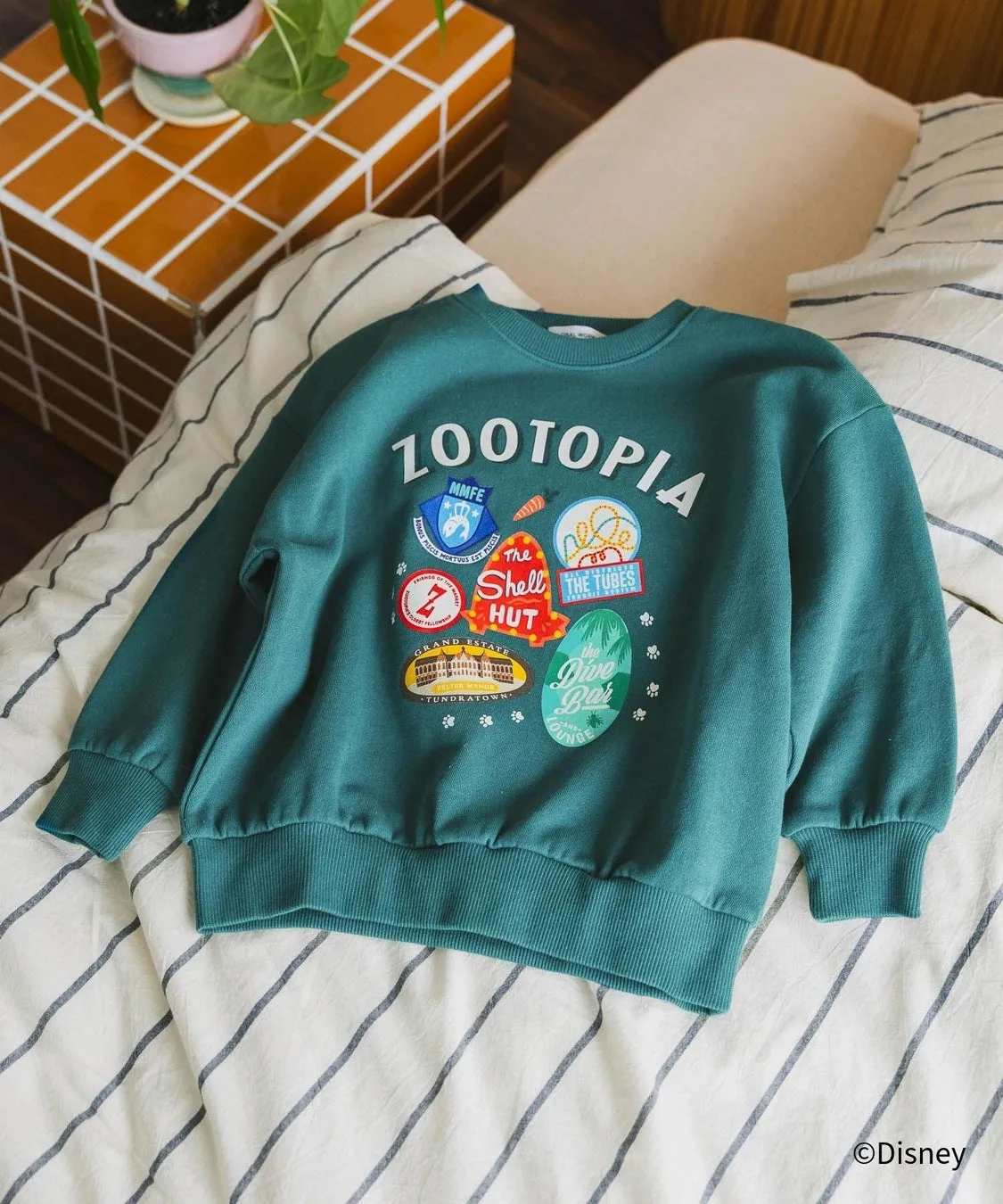 緑色のZootopiaのトレーナーがベッドの上に置かれた写真です。トレーナーには、Zootopiaのロゴと、映画に登場する場所のイラストが描かれています。