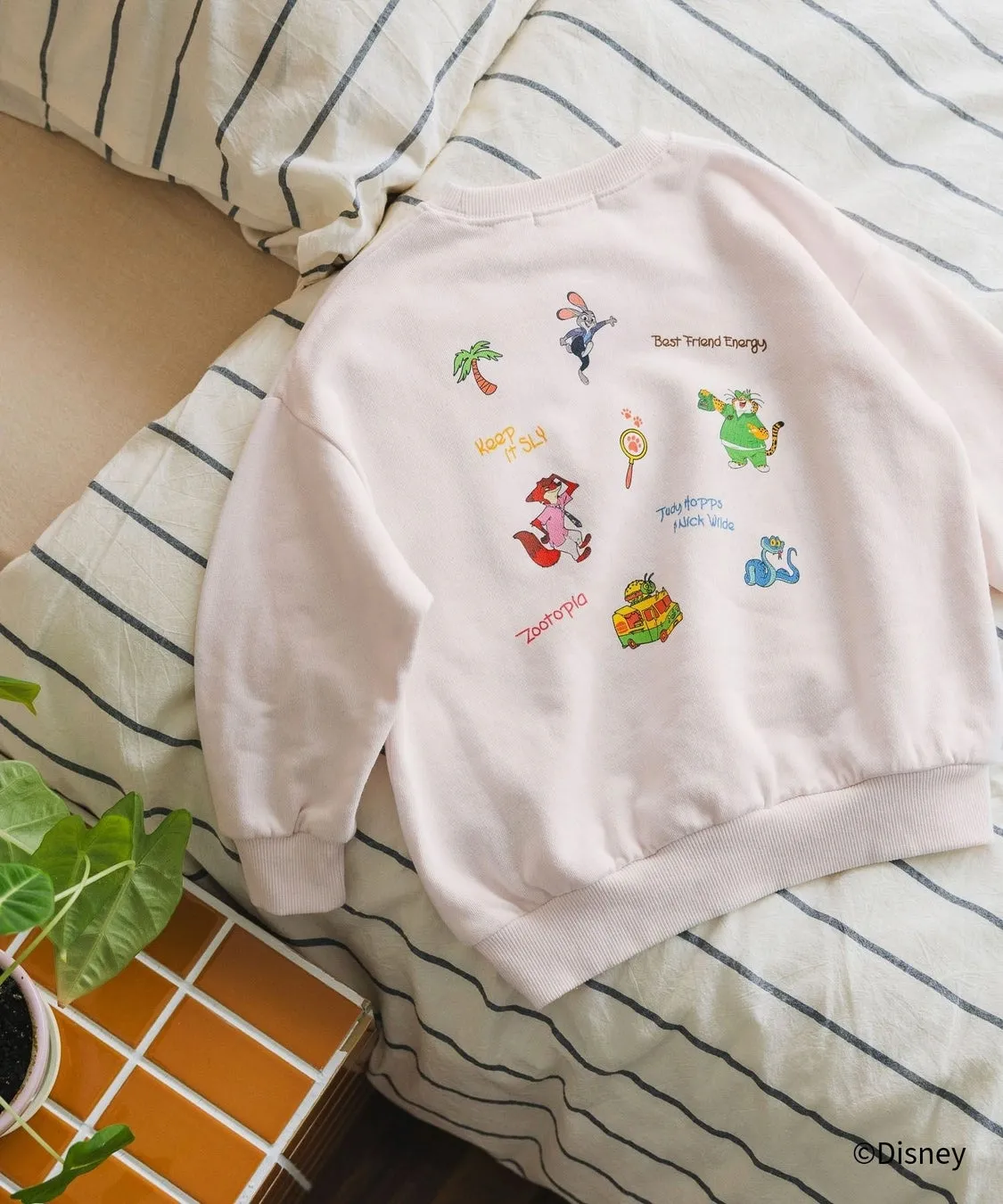 ピンク色のトレーナーの背面を写した写真。トレーナーには、映画「ズートピア」のキャラクターのイラストと英語の文字がプリントされている。トレーナーは白いシーツの上に置かれており、近くには観葉植物とタイル張りのテーブルが見える。