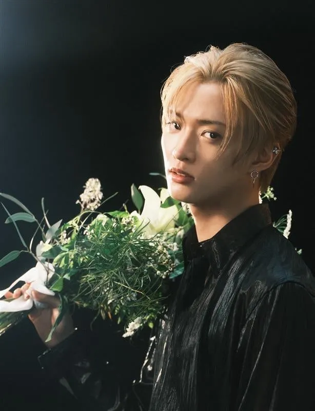 金髪の男性が花束を持ち、こちらを見ている写真。黒いシャツを着ており、背景は黒。