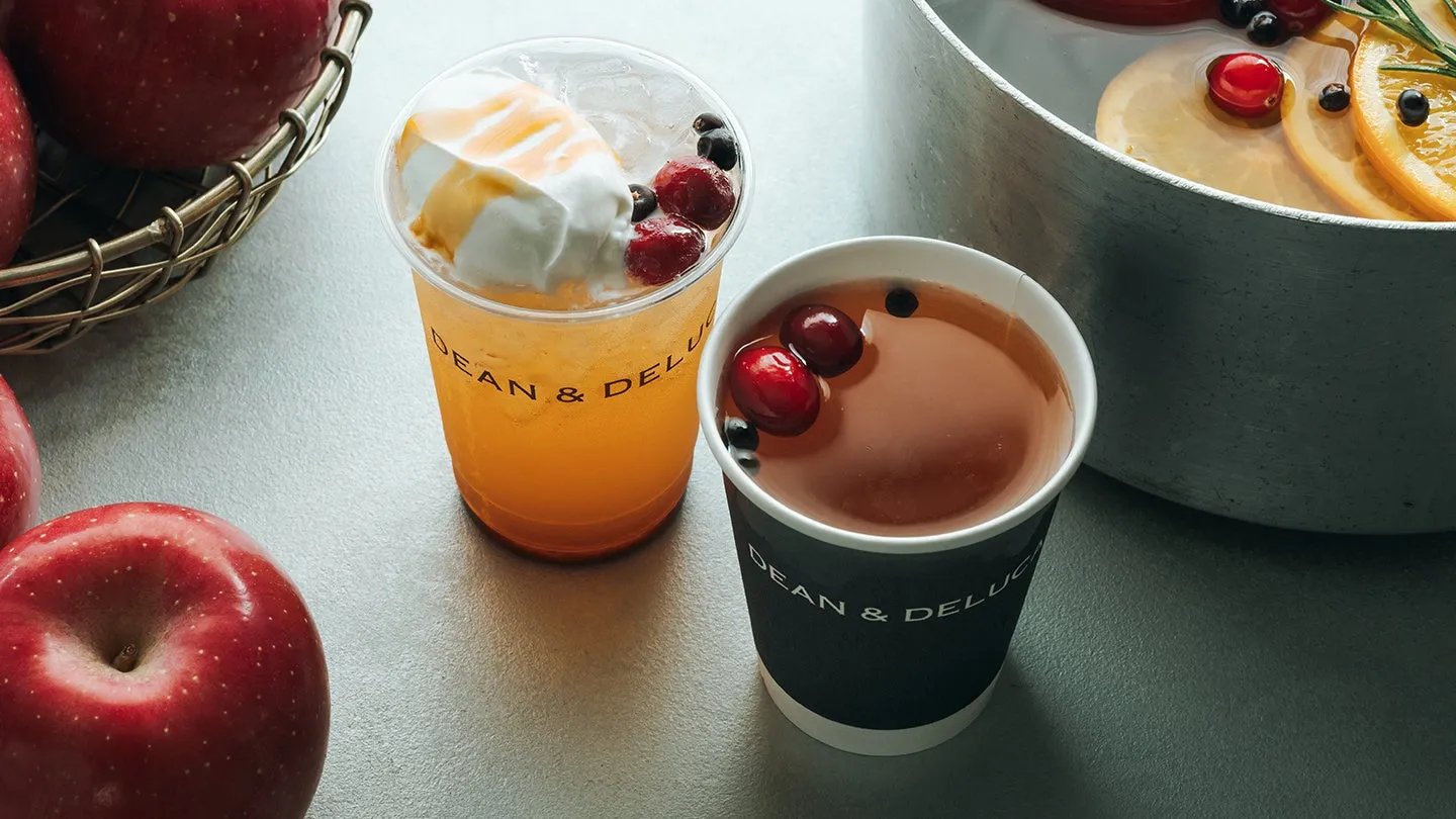 リンゴとドリンクの美味しそうな写真。手前には2つのカップがあり、一つはオレンジ色の飲み物にクリームとベリーがトッピングされ、もう一つは温かい飲み物でベリーが浮かんでいる。背景にはリンゴとフルーツが浮かんだ容器がある。