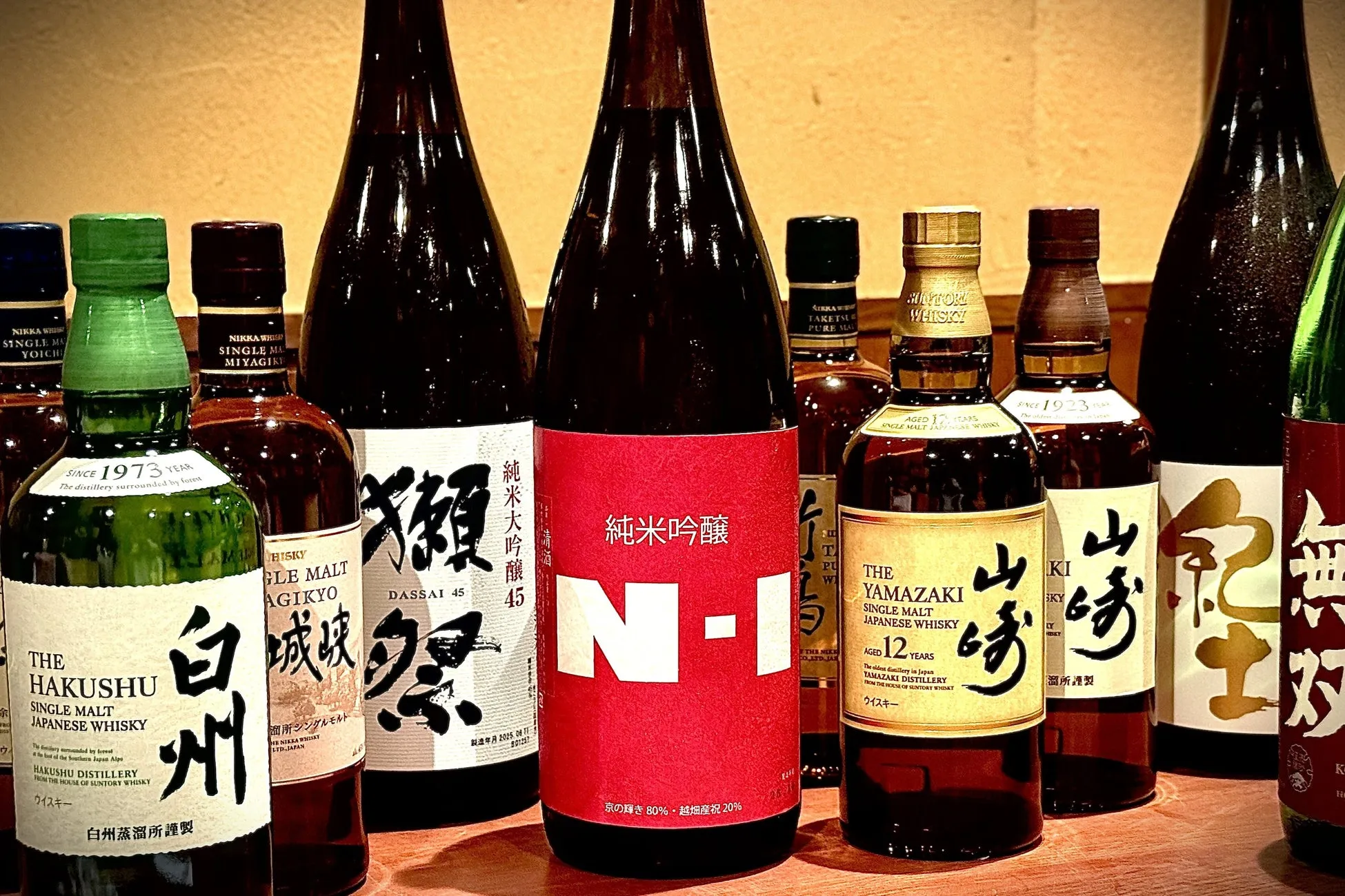 様々な種類の日本酒とウイスキーの瓶が並んでいる写真。ラベルには「白州」、「山崎」、「獺祭」などの文字が見える。