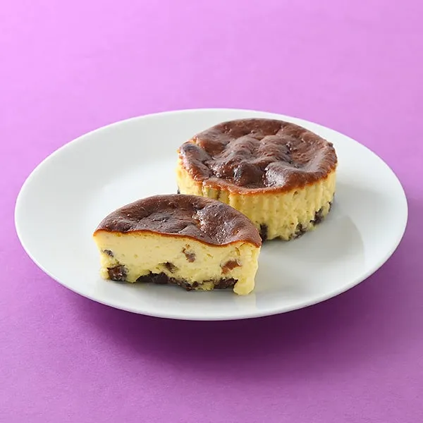 紫色の背景に白い皿に載ったチーズケーキの写真です。チーズケーキは2つあり、1つは半分にカットされています。チーズケーキは焼き目がついていて、中にはレーズンが入っているようです。