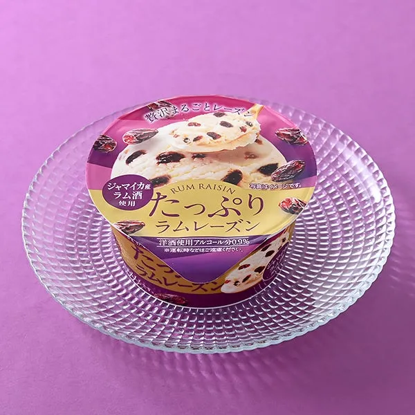 ラムレーズンアイスクリームのカップがガラスの皿に乗っている写真。紫色の背景。