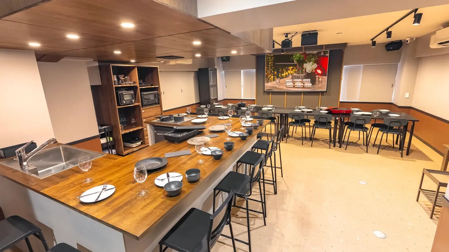 モダンなキッチンとダイニングスペースの写真です。中央に長い木製のカウンターがあり、その周りに黒いスツールが並んでいます。カウンターには、食器、調理器具、グラスがセットされています。奥には、テーブルと椅子が並んだダイニングエリアがあり、壁には絵が飾られています。キッチンには、ステンレス製のシンク、オーブン、棚があります。