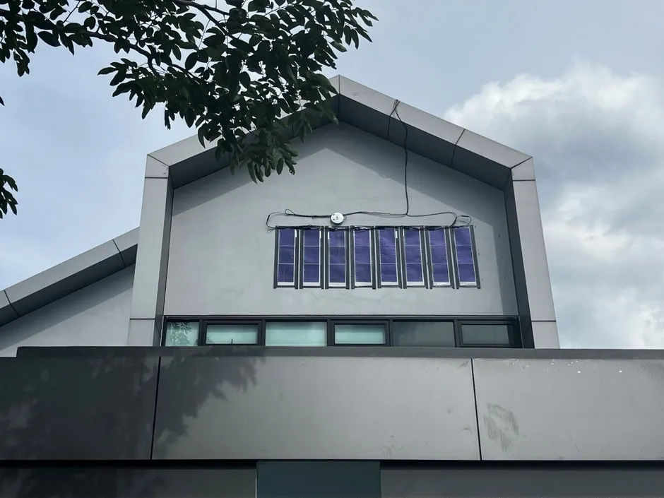 建物の屋根に設置された太陽光パネルの画像です。パネルは窓のように配置されており、建物の外壁はグレーです。空は曇りです。